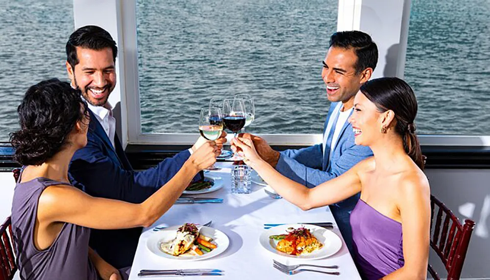 Four people are joyfully toasting with wine glasses at a dining table by the window on a boat
