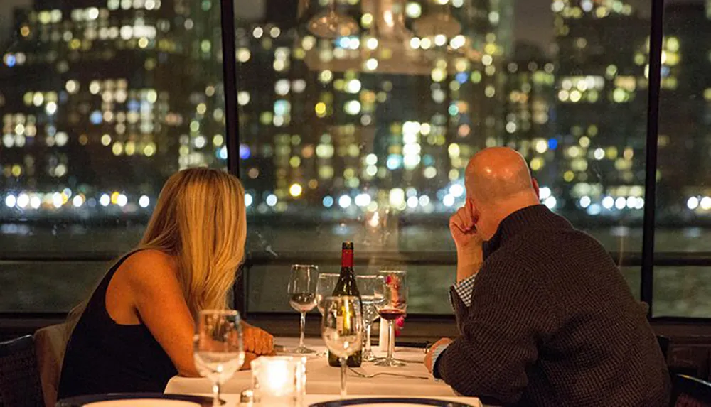 A man and woman sit at a dinner table by a window overlooking a city skyline at night
