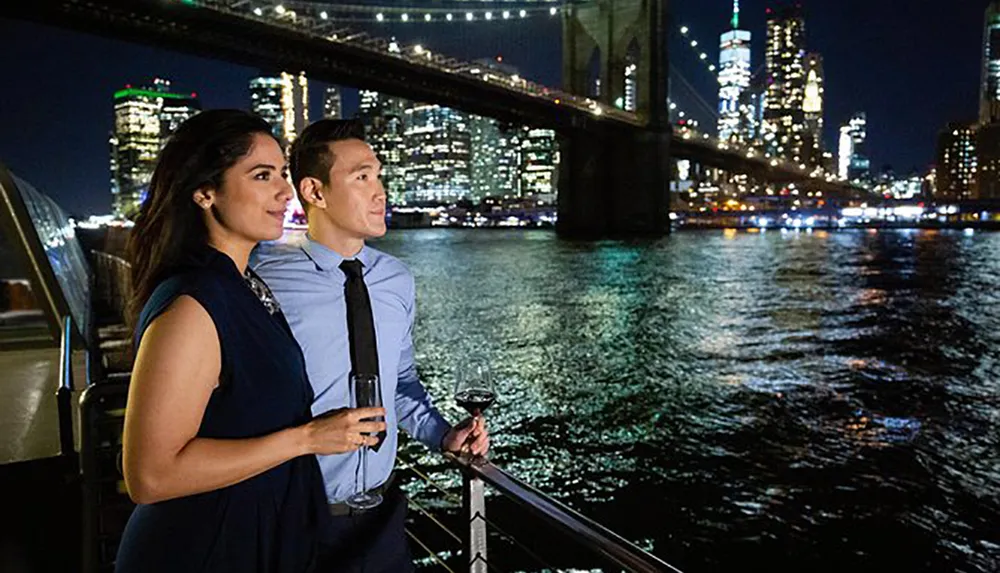 A couple enjoys a nighttime view of a city skyline and a bridge over a river holding wine glasses on a boat or waterfront
