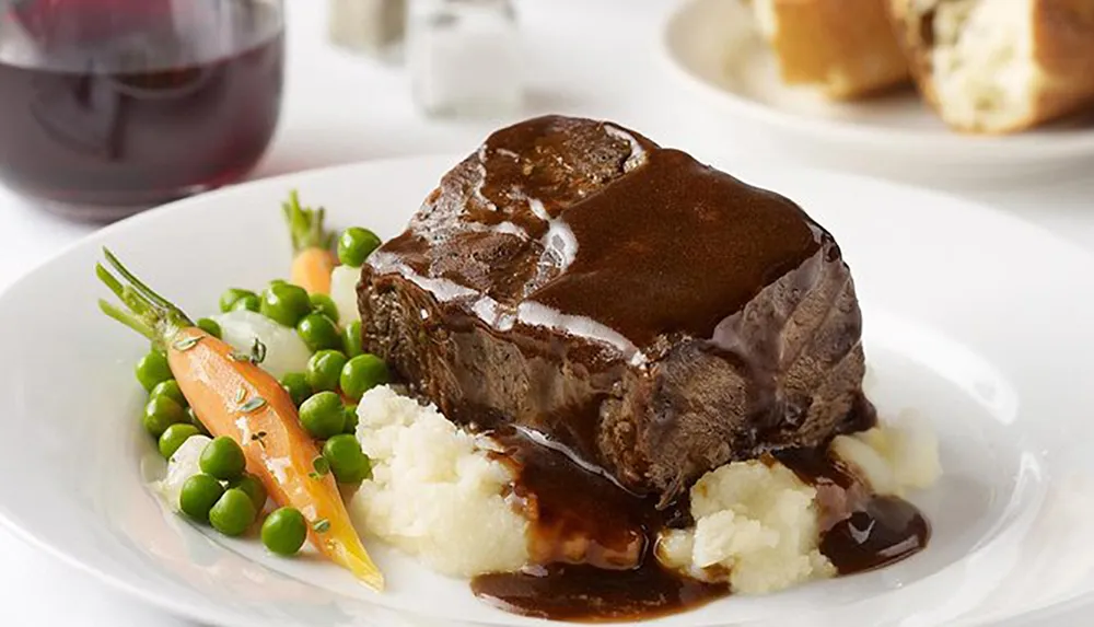 A plate of braised beef with gravy served with mashed potatoes green peas and whole baby carrots accompanied by a glass of red wine in the background