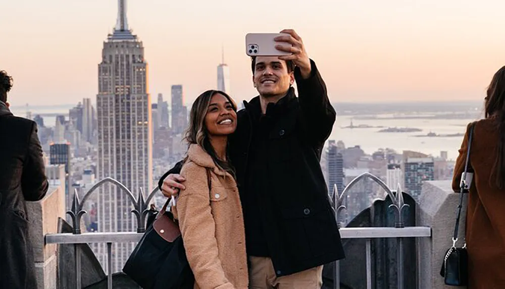 Two people are taking a selfie with a smartphone in front of a scenic urban skyline at dusk