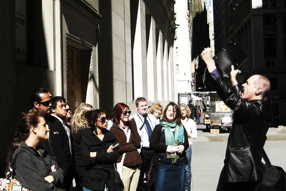 A group of people listens attentively as a man in a black jacket animatedly speaks or performs possibly on a city street or during a walking tour