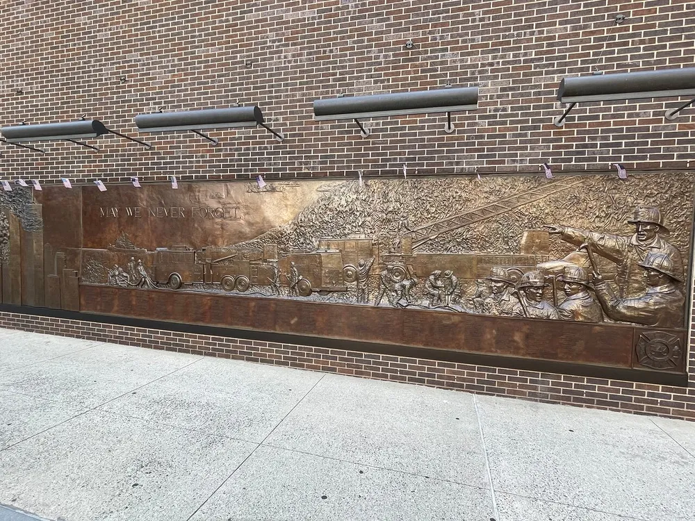 The image shows a bronze relief memorial wall with the phrase MAY WE NEVER FORGET inscribed depicting firefighters in action mounted on a brick wall beneath some hanging purple ribbons