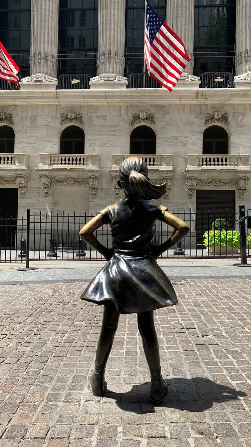 The image shows the Fearless Girl statue facing the New York Stock Exchange framed by two American flags