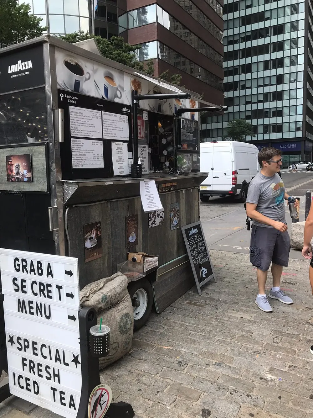 A person stands next to a compact urban coffee cart advertising a secret menu and specialty fresh iced tea