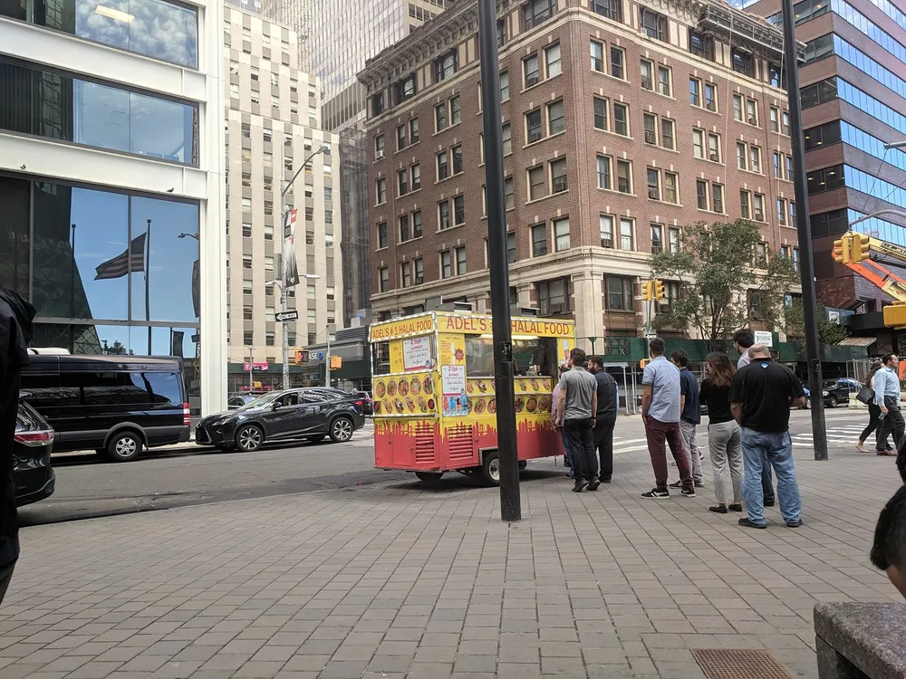 People are queuing at a halal food cart on a bustling city street with surrounding buildings reflecting the diverse architectural styles of an urban environment