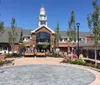 The image shows a sunny day at a well-kept outdoor shopping center featuring a building with a prominent clock tower surrounded by shoppers enjoying the day