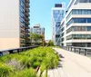 A modern urban park with wooden benches and walkways is nestled between contemporary apartment buildings on a sunny day
