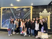 A group of smiling people are posing for a photo in front of a large promotional poster for a theatrical performance.