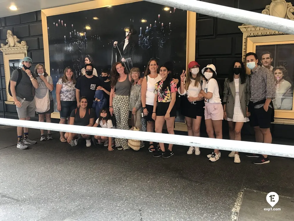 A diverse group of individuals posing for a photo with some wearing face masks at what appears to be an outdoor location with a large decorated frame in the background