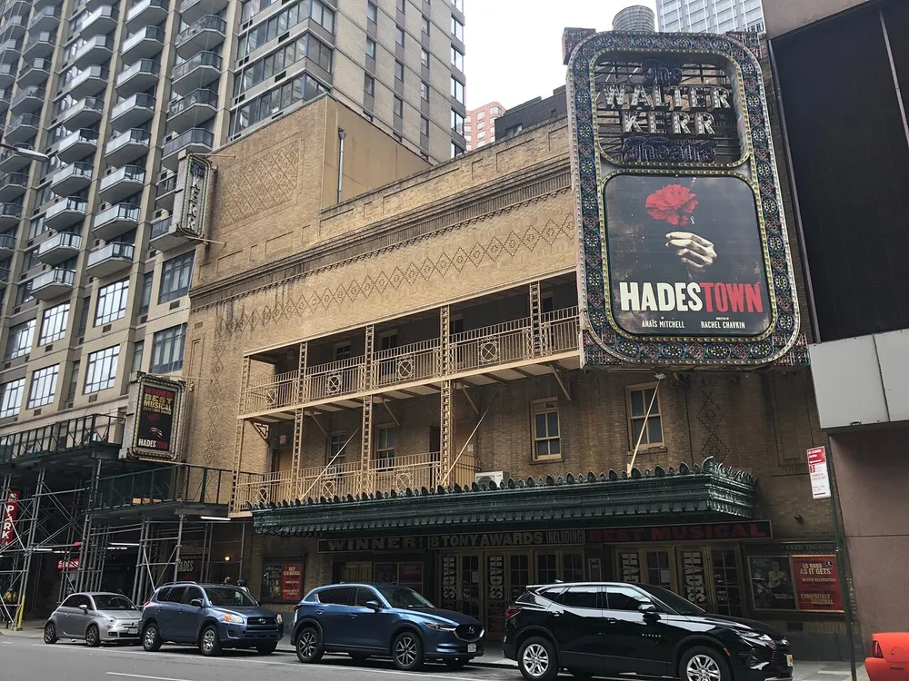 The image shows the exterior of the Walter Kerr Theatre advertising the musical Hadestown on its marquee with cars parked in front and a modern building towering in the background