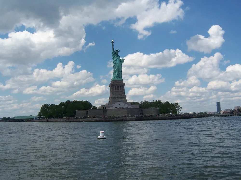 The image shows the Statue of Liberty on its island under a partly cloudy sky as seen from a distance over the water with a buoy visible in the foreground