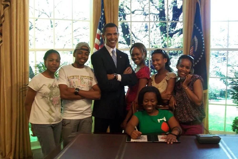 A group of people poses with smiles around a desk in an office-like setting with one person mockingly signing a document and another pretending to take a phone call