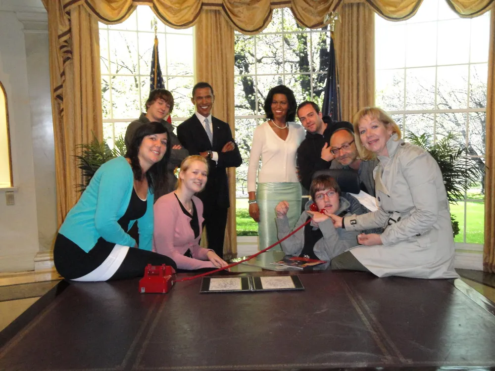 A group of people is posing for a photo with life-sized cardboard cutouts of Barack and Michelle Obama in an elegant room with large windows and draperies