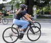 A person wearing a helmet and sunglasses is riding a road bike on a street alongside other cyclists in casual attire