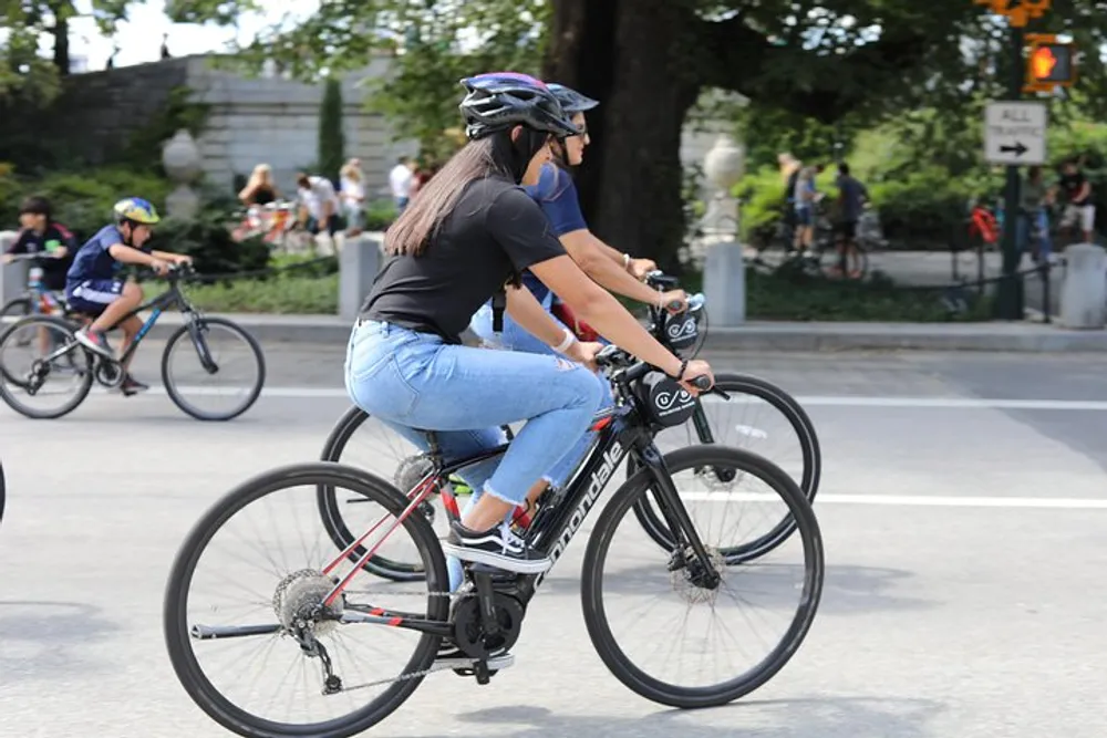 A person wearing a helmet and sunglasses is riding a road bike on a street alongside other cyclists in casual attire