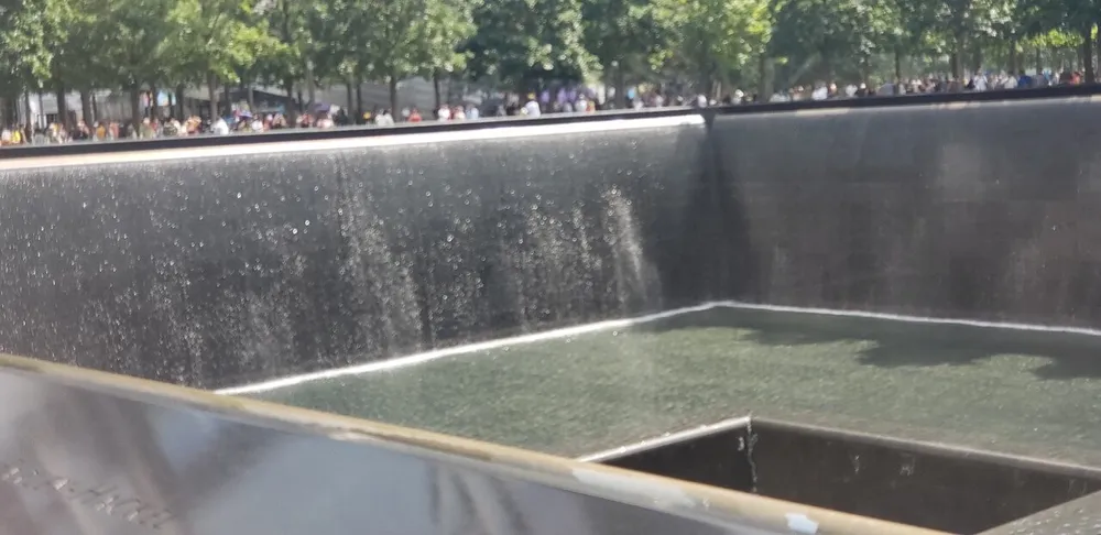 This image captures a large square-shaped fountain with water cascading down its sides surrounded by numerous visitors in the background