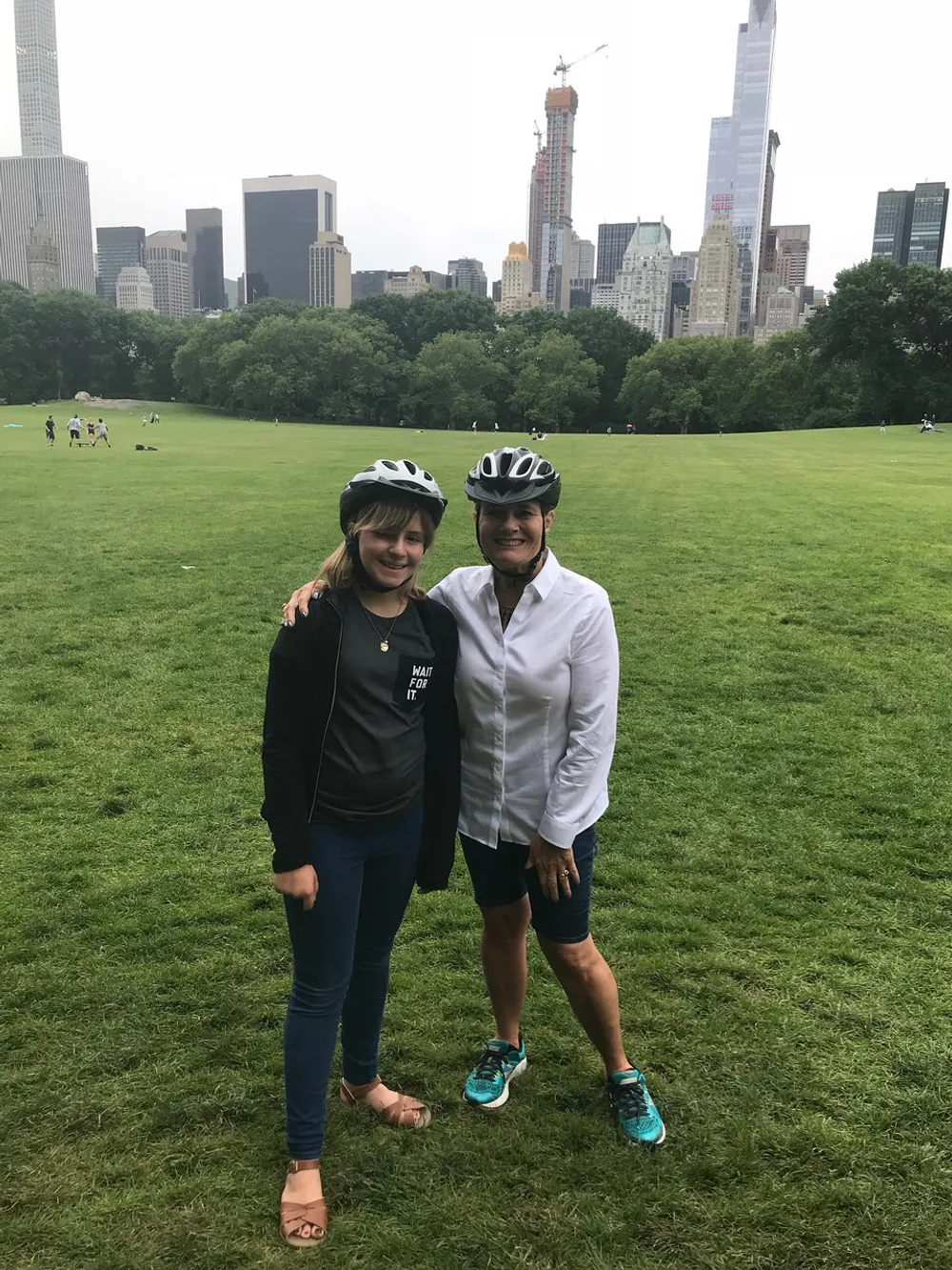 Two people wearing bike helmets are standing together in a grassy park with a city skyline in the background