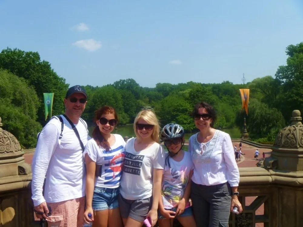 A group of five people is posing for a photo on a sunny day at an outdoor location likely a park with greenery and a body of water in the background