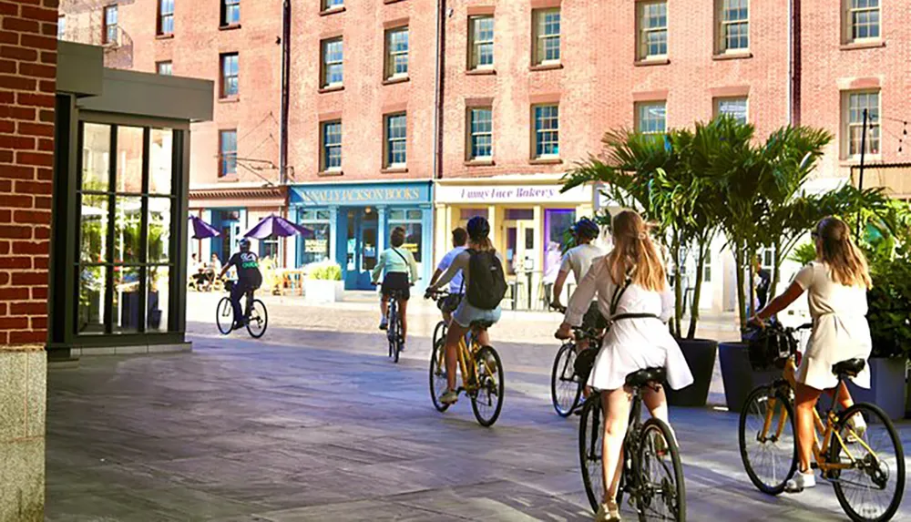 The image shows a group of people bicycling through what appears to be a pedestrian plaza lined with shops and red brick buildings on a sunny day