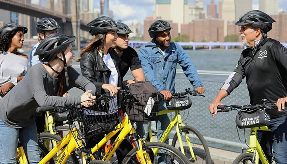 A group of cyclists wearing helmets pauses during a bike tour to listen to their guide with a city skyline and water as the backdrop