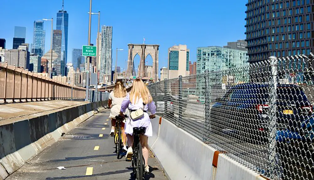 Three individuals are cycling on a bike path with the Brooklyn Bridge and the Manhattan skyline in the background