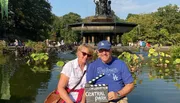 A smiling couple poses in front of a water fountain surrounded by greenery in Central Park, holding a sign indicating they are on location in the famous urban park.