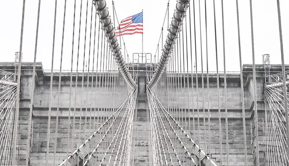 The image shows a symmetrical view of the Brooklyn Bridges cable pattern with an American flag atop the central tower