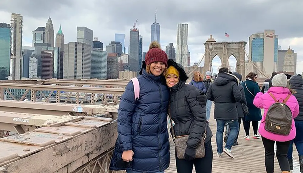 Two people are smiling for a photo on a crowded bridge with a city skyline in the background