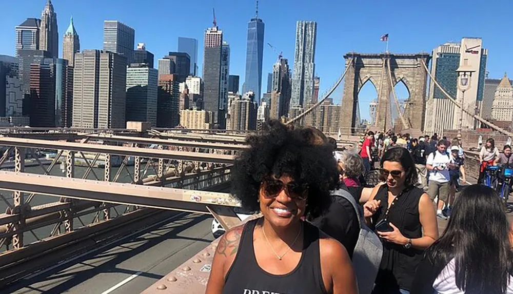 A person is smiling for the camera on a sunny day with the Brooklyn Bridge and the New York City skyline in the background