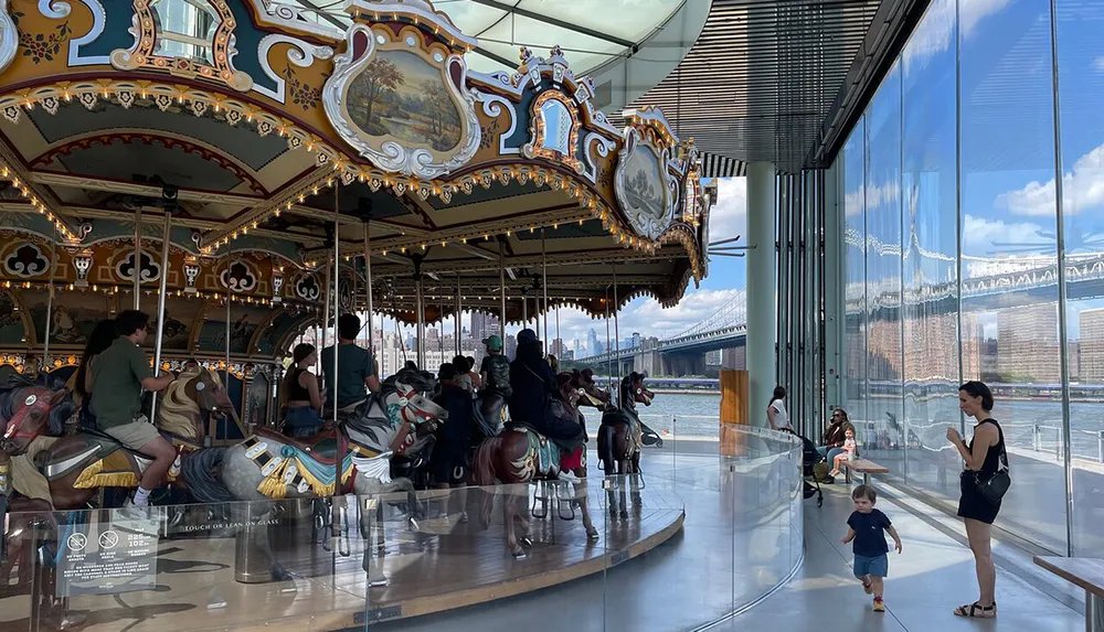 A classic carousel with ornately decorated horses is enjoyed by visitors inside a modern glass-enclosed pavilion overlooking a bridge and waterfront