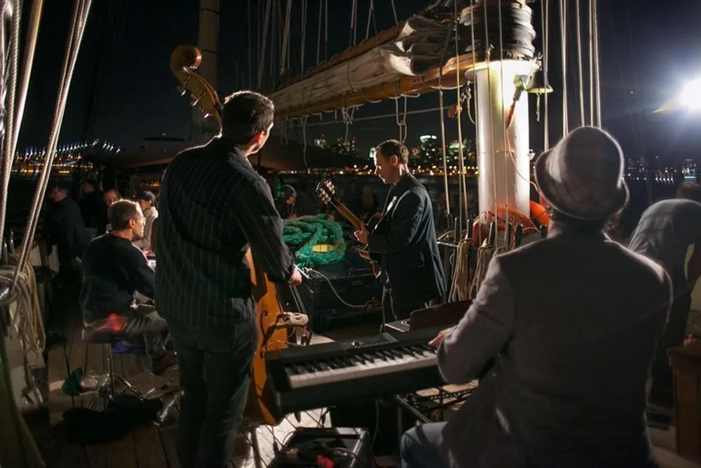 A group of musicians is performing on the deck of a boat at night with city lights in the background