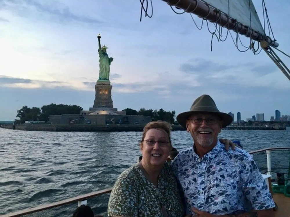 Two smiling individuals are posing for a photo on a boat with the Statue of Liberty in the background during what appears to be dusk