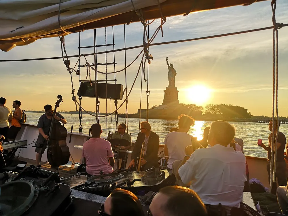 A group of people enjoys a musical performance on a sailboat with the Statue of Liberty in the background during sunset