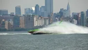 A high-speed boat is creating a large spray of water as it cuts through the waves in front of a cityscape.