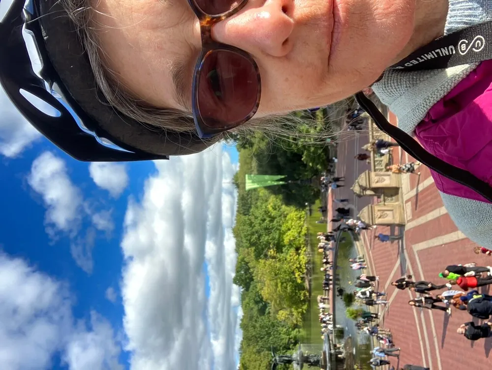 A person wearing sunglasses and a bike helmet takes a selfie with a vibrant partly cloudy sky above and a bustling public place with people and a fountain in the background with the image rotated 90 degrees to the right