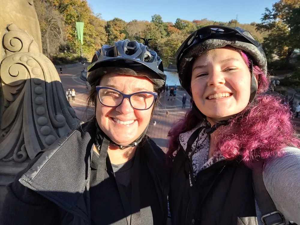 Two people wearing bike helmets are smiling for a selfie with a sunny park scene in the background