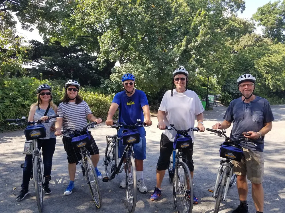 Five individuals wearing helmets and casual attire are standing with their bicycles seemingly ready for a group ride in a leafy outdoor setting