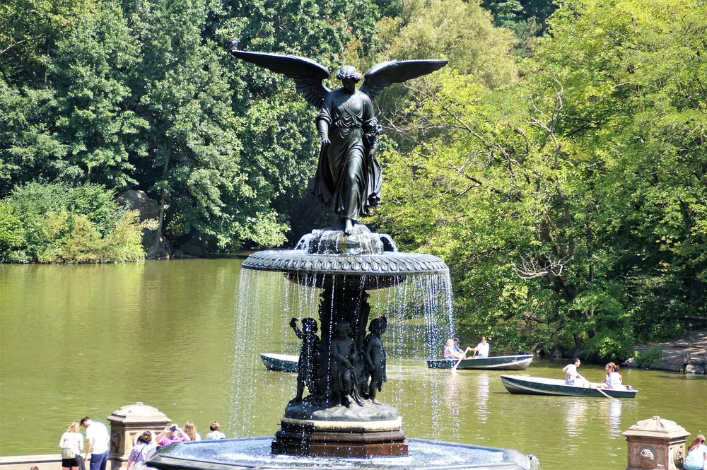 The image shows the Angel of the Waters statue atop the Bethesda Fountain in Central Park New York with people rowing boats in the background