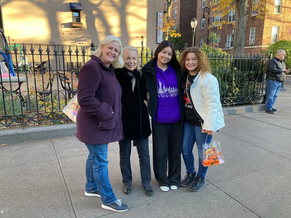 This image shows four people smiling at the camera on a sunny day on a city sidewalk holding what appears to be trick-or-treat bags suggesting they may have participated in a Halloween-oriented event