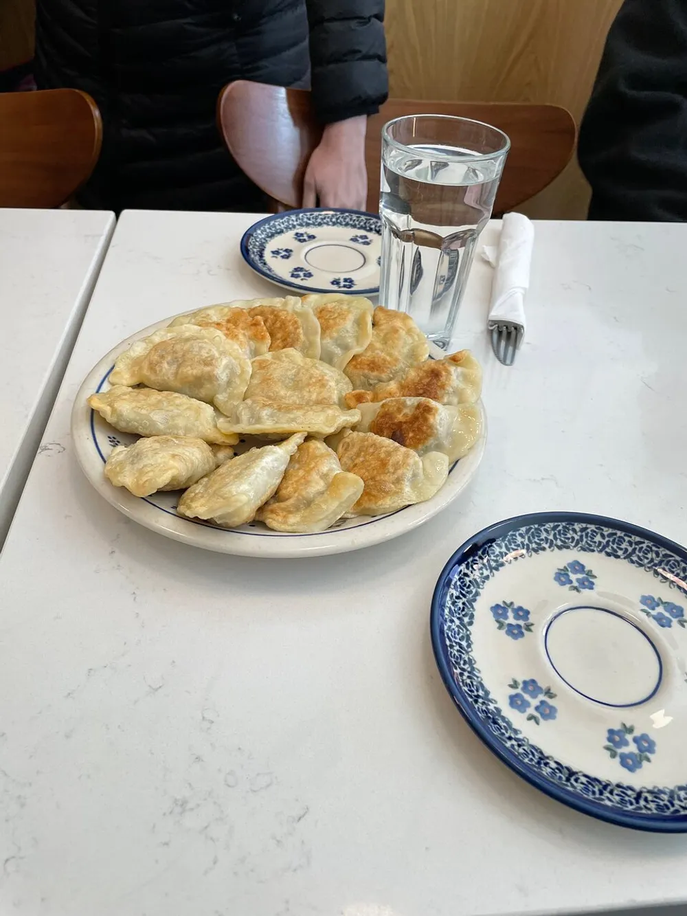 A plate of golden-brown dumplings is served on a table in a casual dining setting accompanied by a glass of water and decorative empty plates with a person seated across the table
