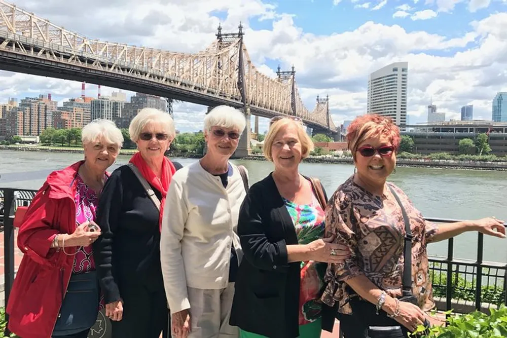 A group of five joyful women pose together in front of a bridge and a city skyline on a sunny day