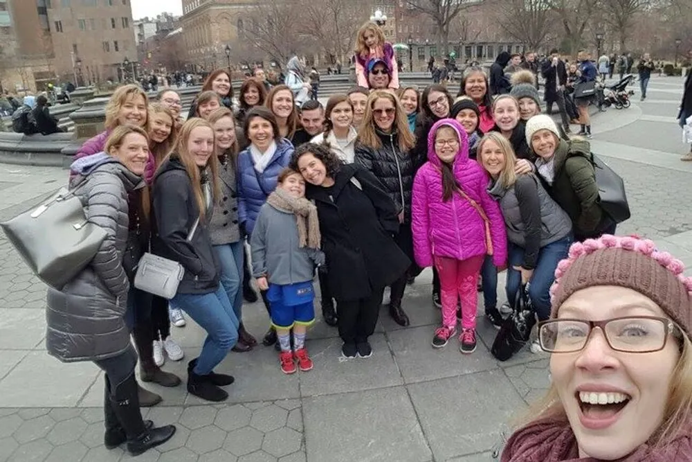 A group of smiling people of various ages are posing for a selfie with one person in the foreground holding the camera and extending her arm to capture everyone