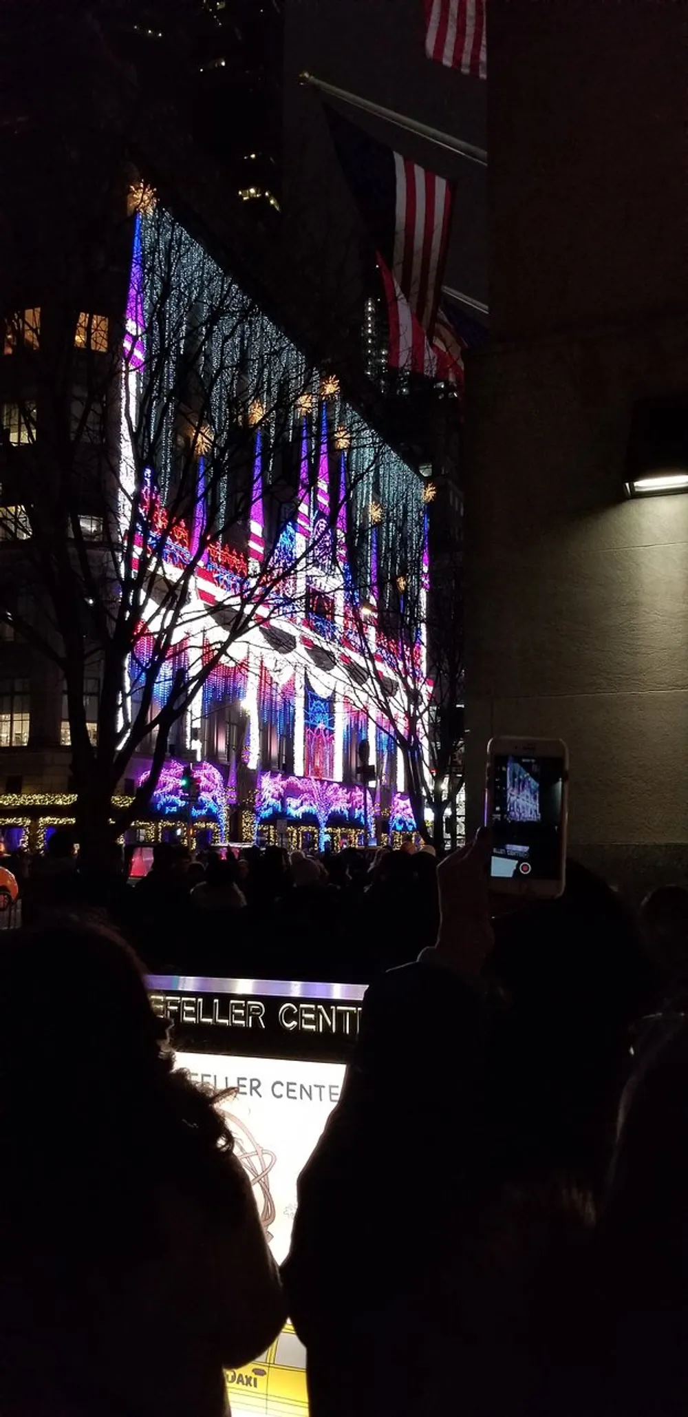 A crowd of people is watching a colorful light display on a building with someone in the foreground taking a photo with their phone and American flags hanging alongside the architecture