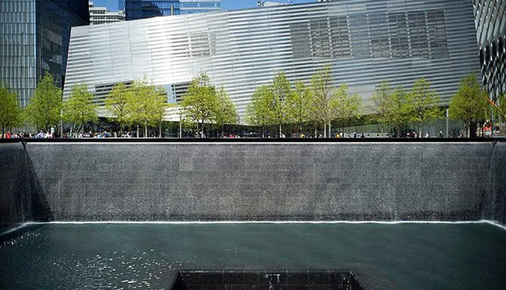 The image shows one of the reflecting pools at the 911 Memorial in New York City with a modern building in the background and young trees lining the area