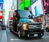 A black limousine is parked on a bustling street with bright billboards and a yellow taxi passing in the background likely depicting a scene from Times Square in New York City