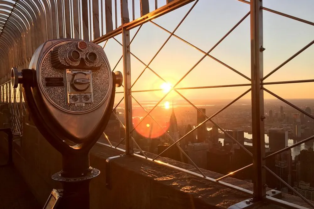 The image captures a coin-operated binocular viewer overlooking a panoramic cityscape bathed in the warm glow of a setting sun