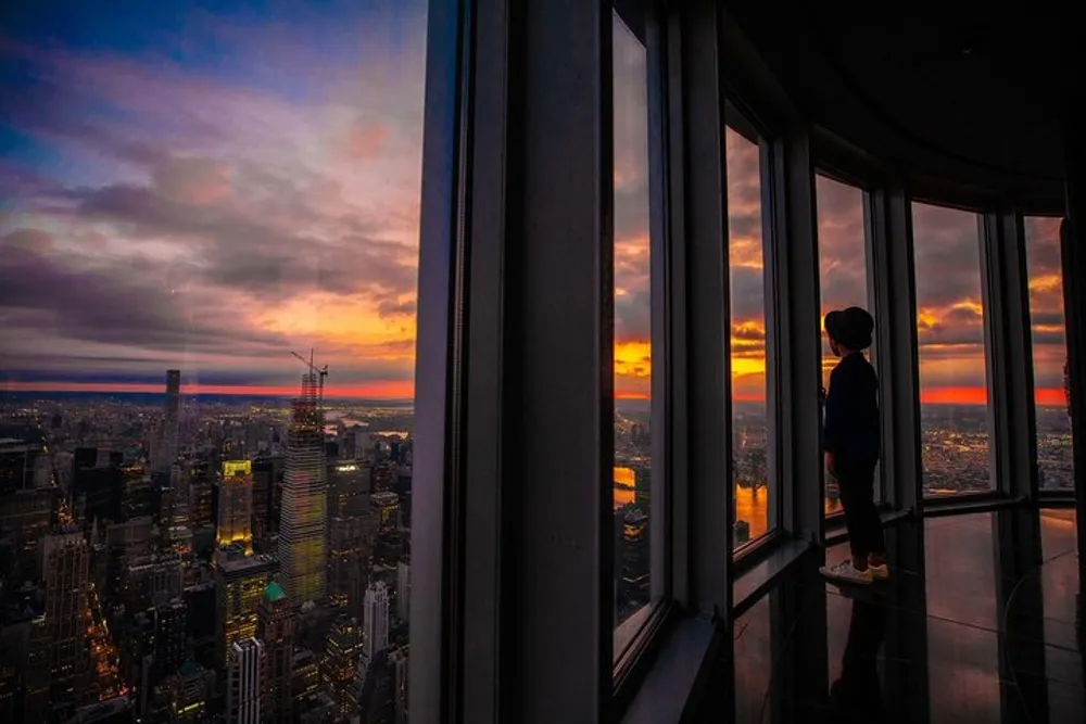 A person stands in a high-rise building gazing out at a sweeping cityscape under a vibrant sunset