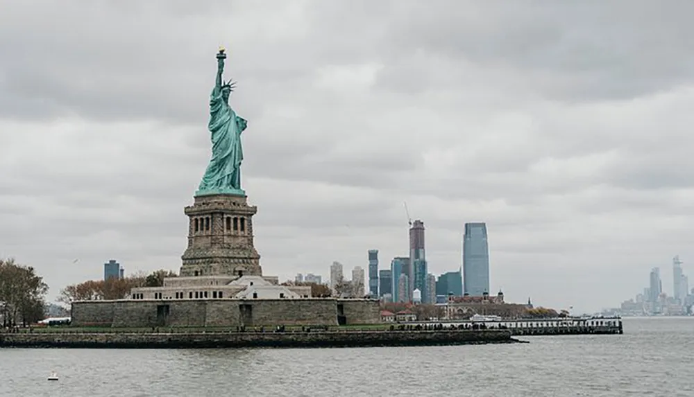 The Statue of Liberty stands in the foreground with an overcast sky overlooking the water with New York Citys skyline in the distance