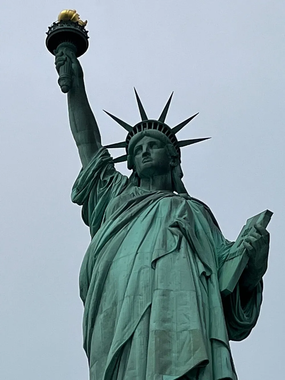 The image is a close-up of the Statue of Liberty against a clear sky highlighting the iconic torch and crown
